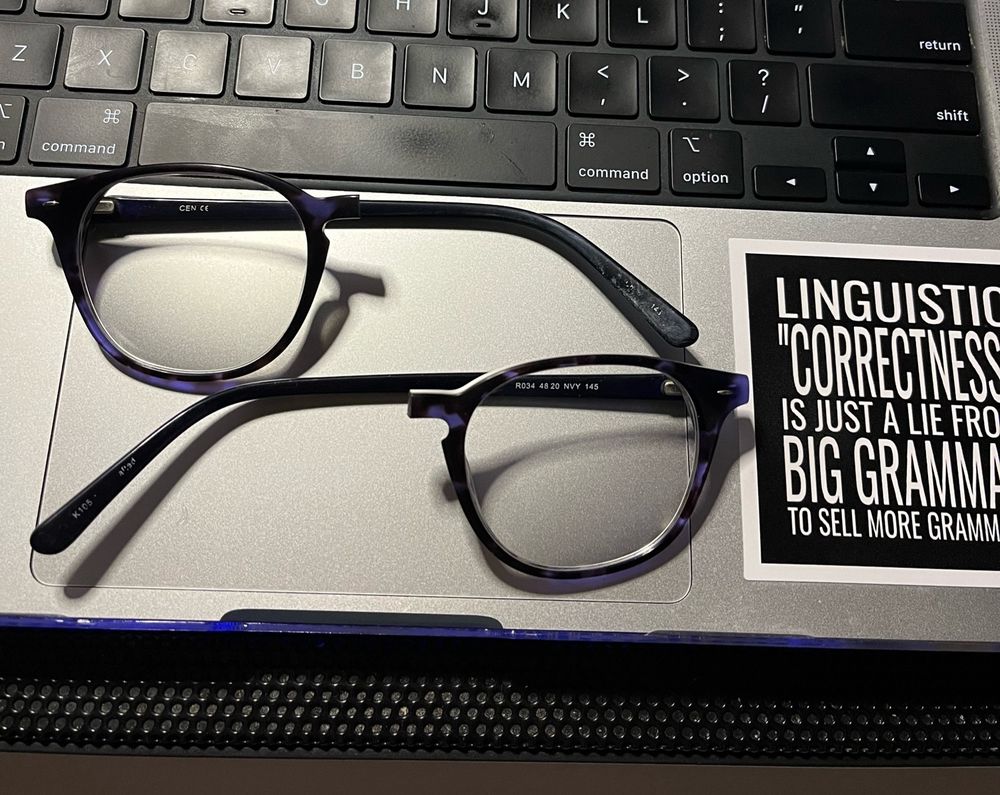 A pair of glasses sitting on a MacBook touchpad. The glasses are aligned strangely because the nose bridge has snapped in half. Next to the glasses is a @lingthusiasm sticker reading “Linguistic correctness is just a lie from Big Grammar to sell more grammars.”