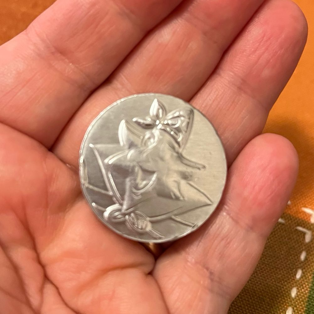Closeup of a small silver-foil-covered chocolate coin in someone’s hand. Embossed on the coin is a stingray? starfish? With a pirate-style bandanna and an eye patch.