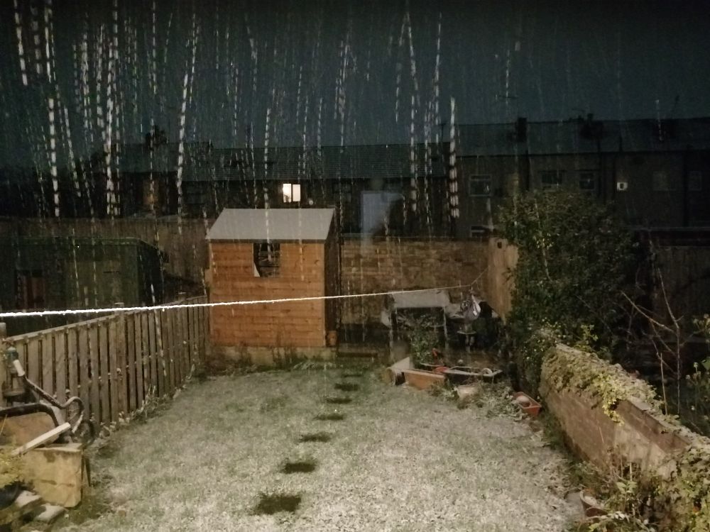 Somewhat fuzzy picture of a small back garden with terraced houses behind, taken at night, with the outside light on. There is grass and a path of paving stones leading to a raised patio with a small wooden shed and garden benches on it. A washing line crosses the garden. The washing line, grass, shed roof and benches have a light covering of snow. Snow is falling, lit up by the light to show as vertical trails of white dots against the grey sky and dark houses. 