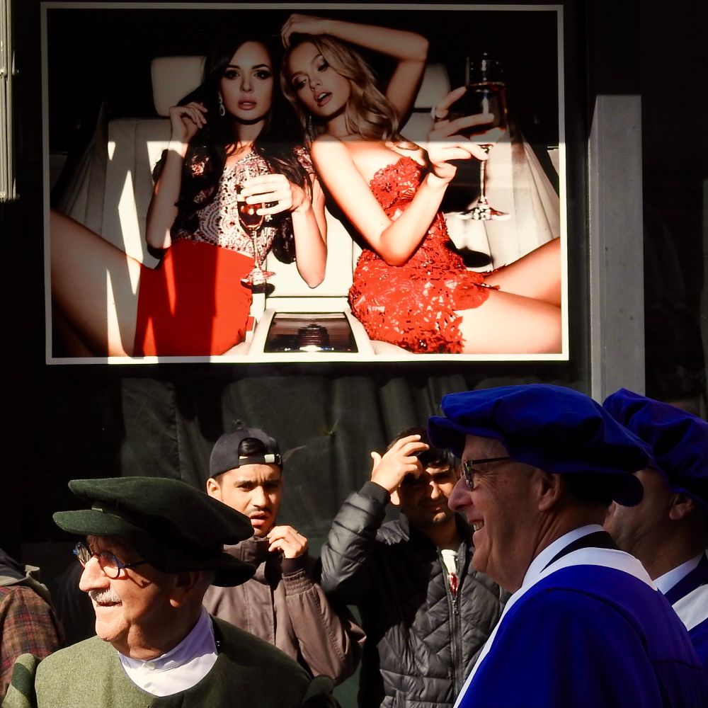 Auf einem großen Werbeplakat oben sitzen zwei elegant gekleidete Frauen in einem Auto und halten Gläser in der Hand. Darunter stehen mehrere Menschen im Freien, darunter ältere Männer in historischen oder festlichen Gewändern sowie jüngere Männer in Alltagskleidung. 