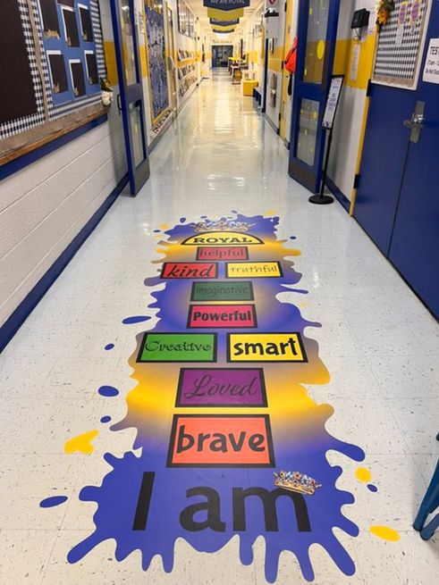 A hopscotch board painted on the floor in a school hallway. In front of the boards, the words "I am" are painted, and the blocks in the board include adjectives such as brave, loved, creative and smart.