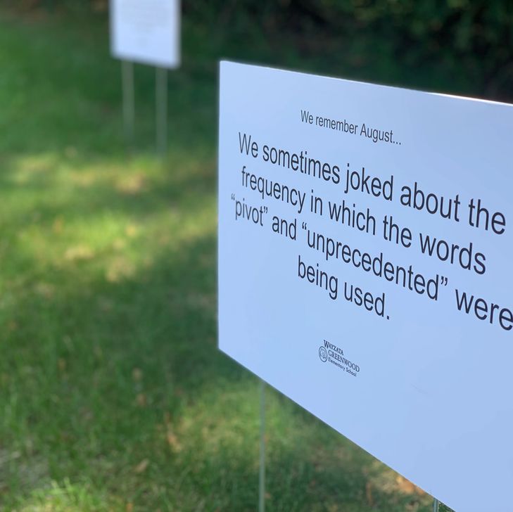 A close-up image of a small white sign stuck in grass. The sign reads "We remember August... We sometimes joked about the frequency in which the words 'pivot' and 'unprecedented' were being used."