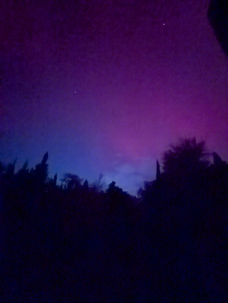 A forested skyline in shadow lines the foreground to a deep cerulean blue sky shifting to a blend of magenta and royal purple from the Aurora Borealis seen over Coastal Maine on October 10th, 2024. 