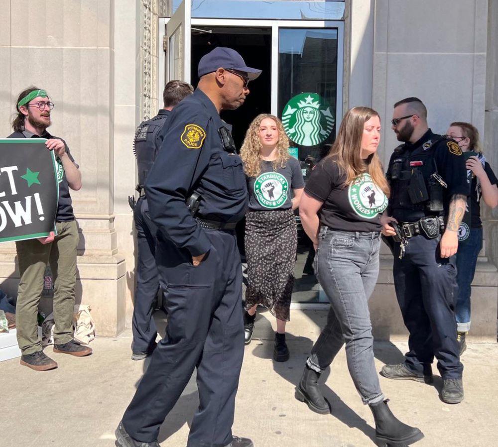 Two baristas in union t-shirts being escorted out of a starbucks location by police with their hands appearing to be tied behind their backs.