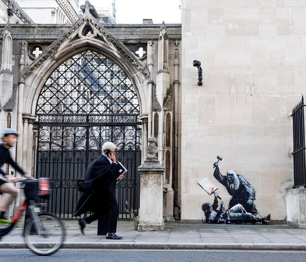Zdjęcie kamiennej ściany Royal Courts of Justice z wyrytym ulicznym dziełem sztuki przedstawiającym sędziego używającego swojego młotka sądowego do uderzenia protestującego leżącego na ziemi z rękami w powietrzu, jedną ręką trzymającego pustą tabliczkę. Na tabliczce znajduje się krew, prawdopodobnie z powodu ataku sędziego. Na pierwszym planie, rozmazany w ruchu, rowerzysta i sędzia w peruce i todze rozmawiają przez telefon.