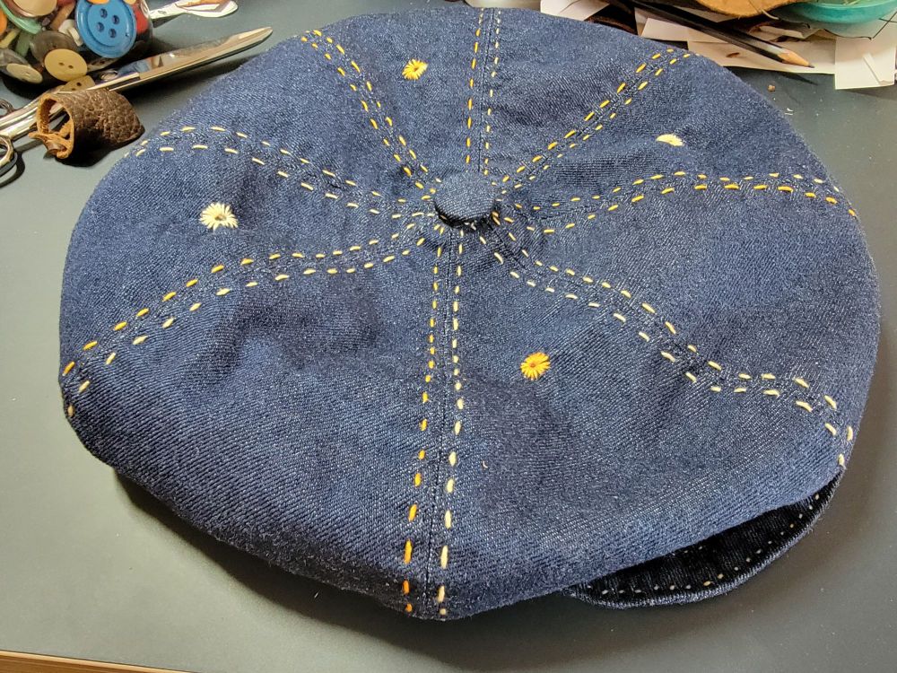 A dark blue denim newsboy cap with variegated yellow stitching. Has four yellow eyelet vents