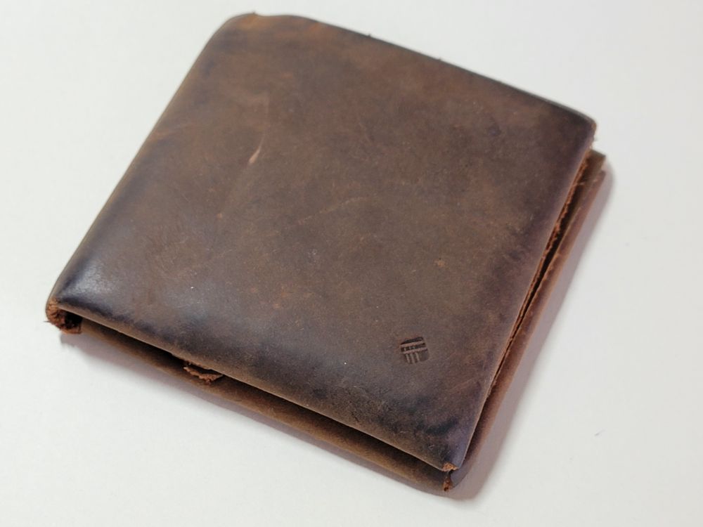 A square brown leather wallet on a white surface. A small logo for the designer is embossed in the bottom right corner.