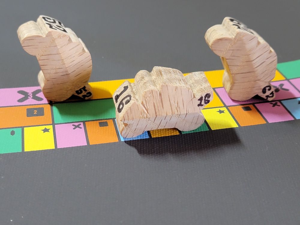 Three hand-carved wooden motorcycle game pieces on a colourful track. Two of the pieces are standing on their tail to represent a wheelie.