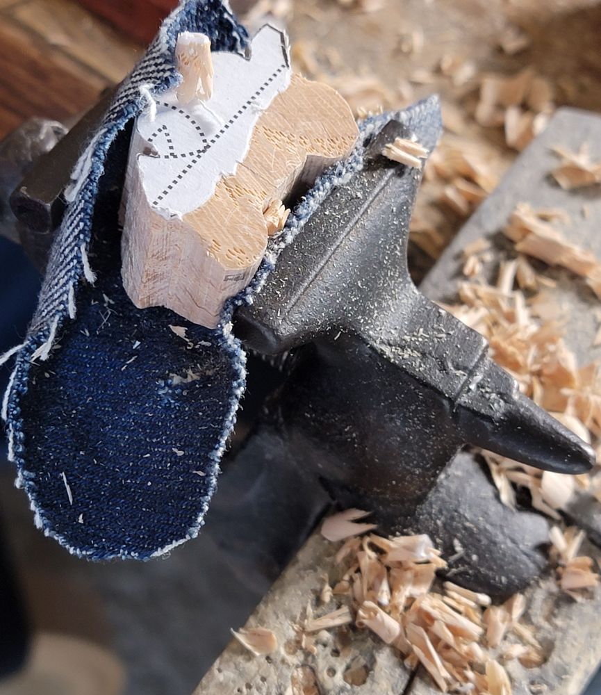 A piece of wood is being carved to resemble a motorcycle with a rider. The wood is clamped in a small antique vice, surrounded by wood shavings.