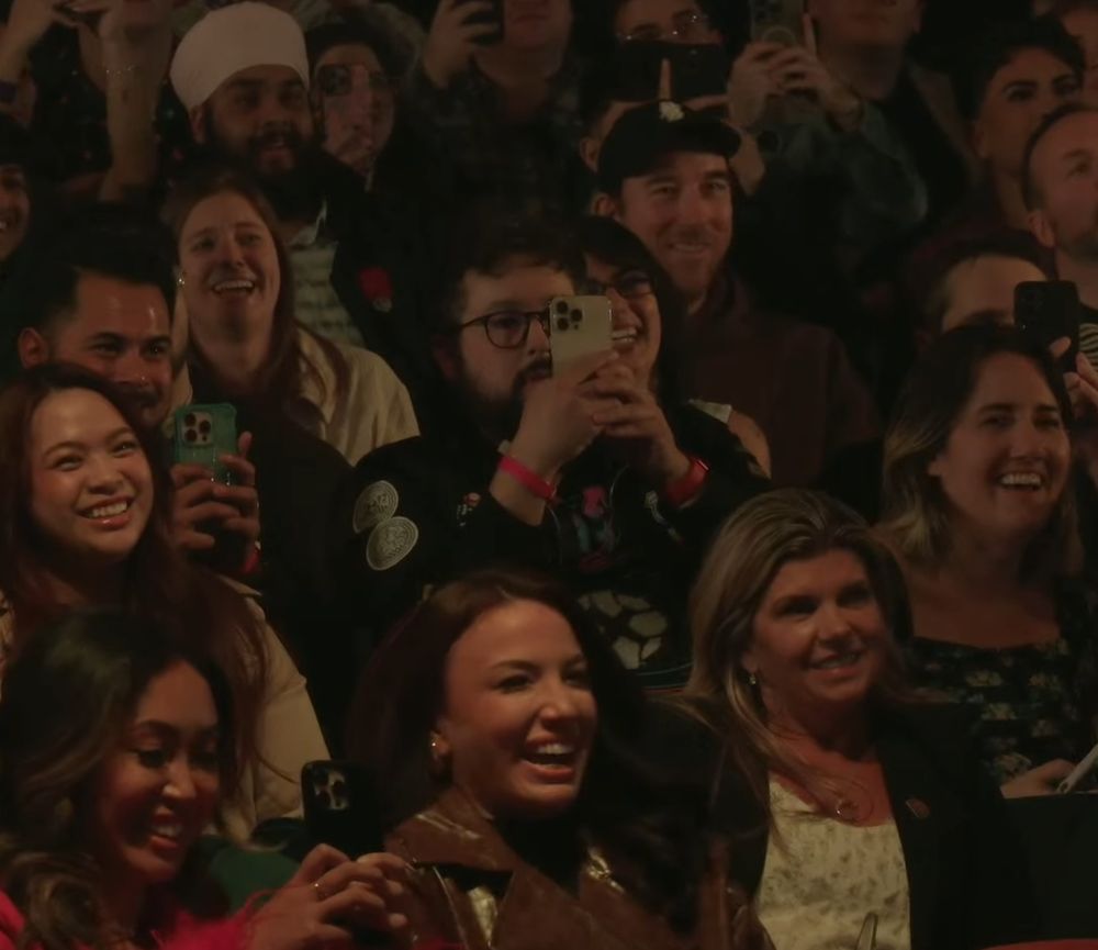IGN's Jeffrey Vega in a crowd at the Taco Bell Live Mas event. Everyone in the crowd is smiling and he looks very stern while holding up his phone to take footage.