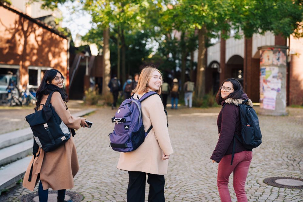 drei Studentinnen laufen in der Herbstsonne über den Campus und schauen über die Schulter in die Kamera