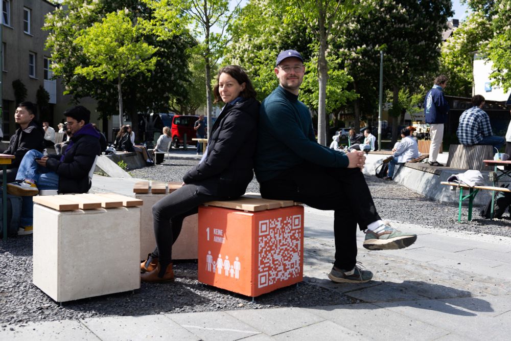Zwei junge Menschen sitzen in Freien Rücken an Rücken auf einem organgefarbenen Würfel, auf dem das Symbol des SDG1 aufgemalt ist.Im Hintergrund Campusleben.  