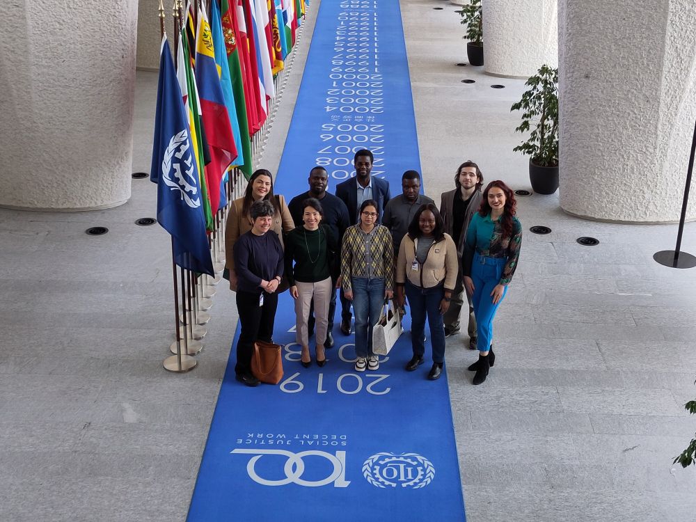 Ten persons standing on a long blue carpet with years next to national flags and smiling into the camera above.