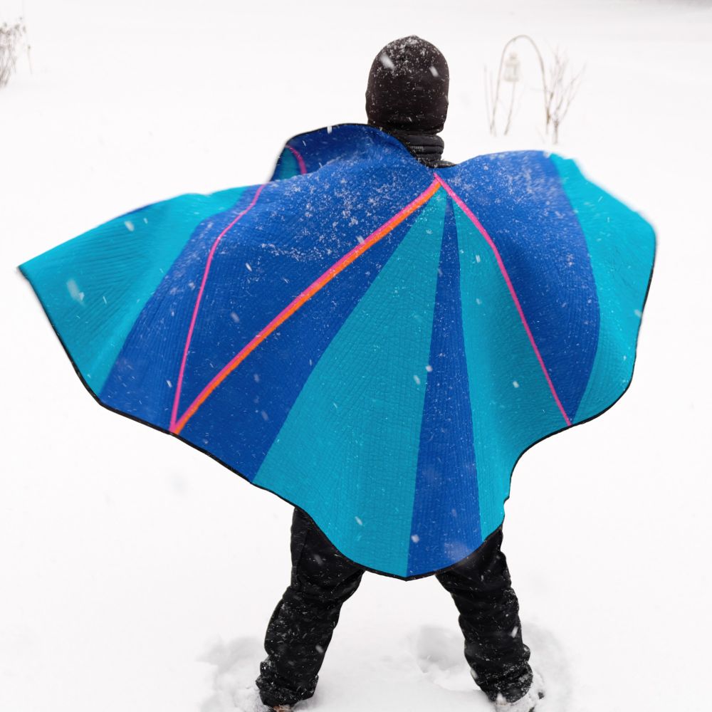 Back view of someone wearing a vibrant geometric quilt like a cape in snowy weather, creating a superhero-like silhouette.
