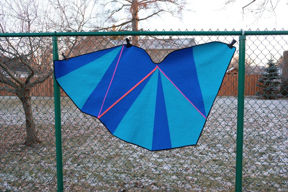 A geometric quilt in blue and turquoise with pink accent lines hanging on a green chain-link fence.