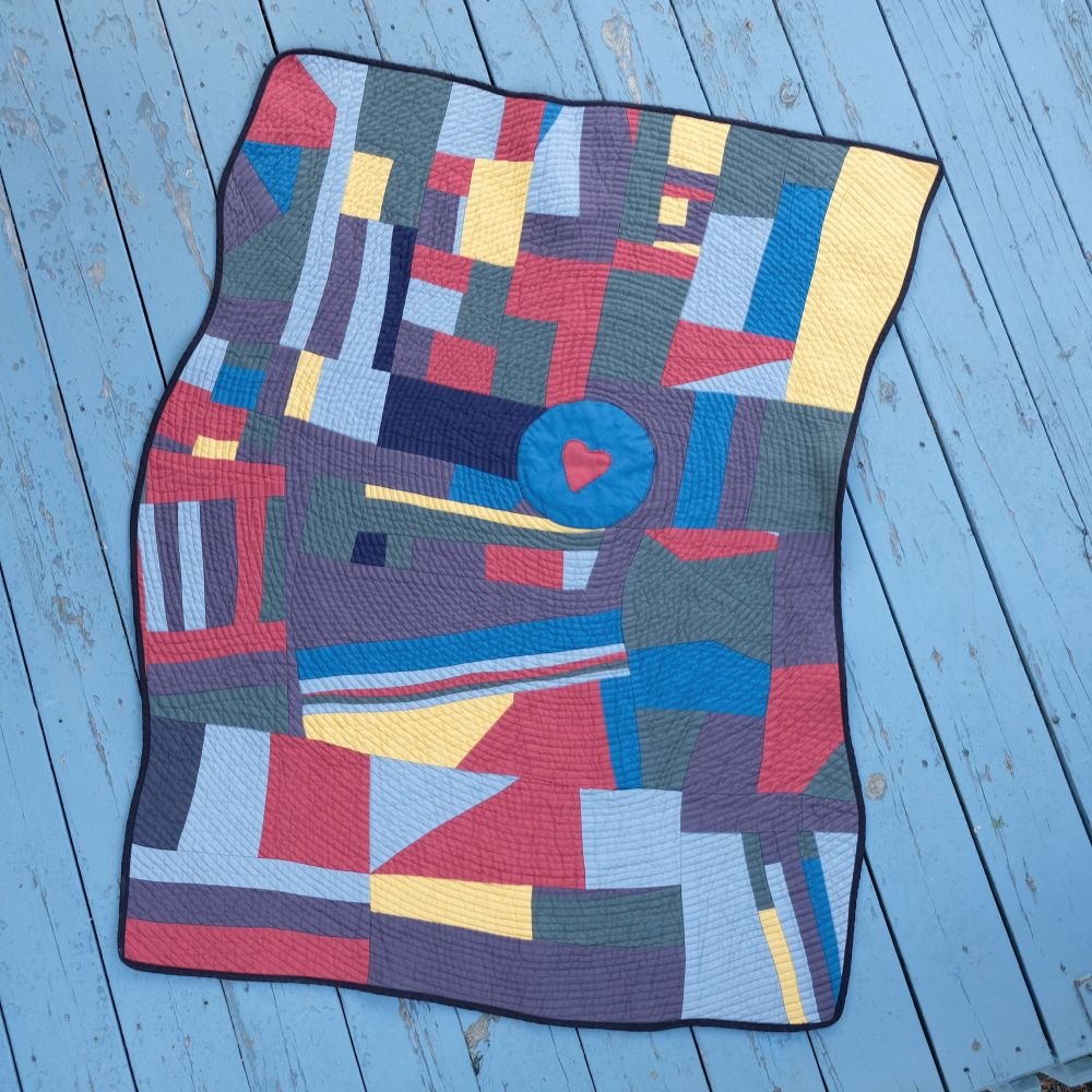 Geometric quilt with red, blue, yellow and gray patches on blue wooden boards. Blue circle with red heart in center. Black binding.