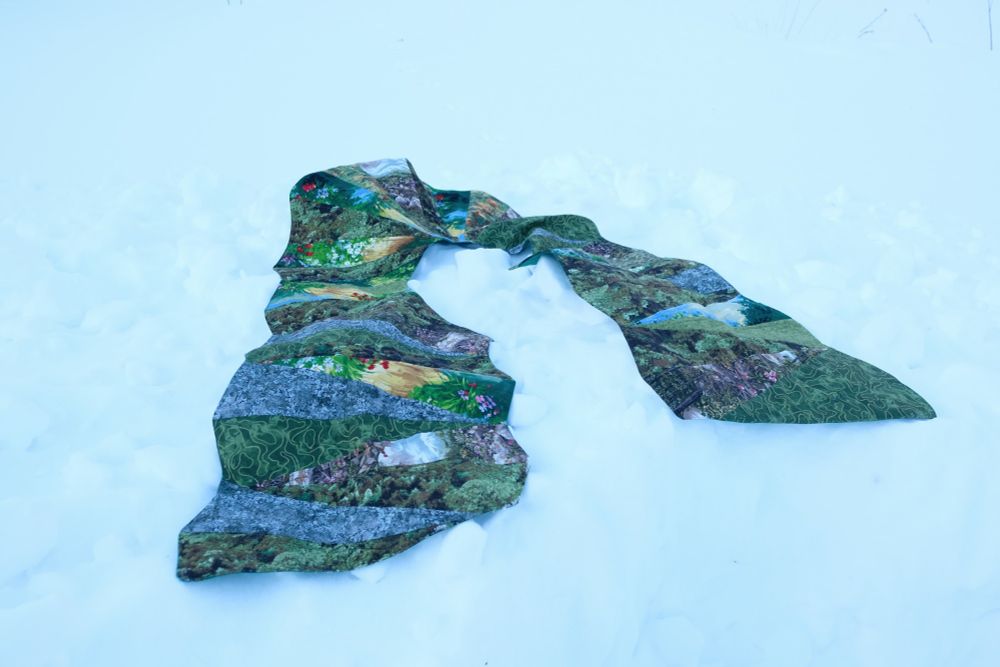 Quilted stole measuring 56 x 77 inches, featuring a landscape design inspired by the green stripe of the Pride flag. The quilt combines various green fabrics in triangular gore shapes showing mountains, meadows, and wildflowers against a snowy white background.