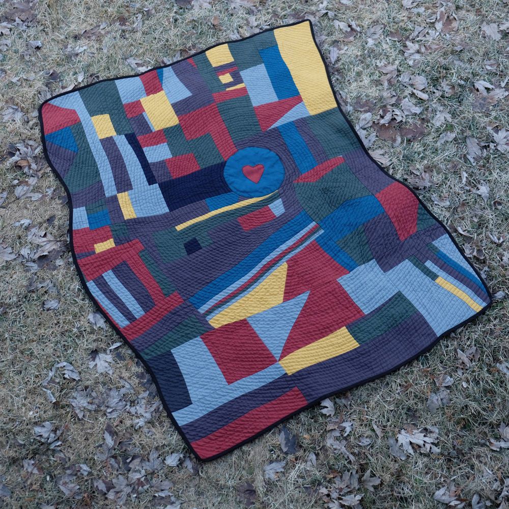 Patchwork quilt on grass and leaves. Made with linen fabric in blue, red, yellow and gray. Features blue circular center with red heart detail.