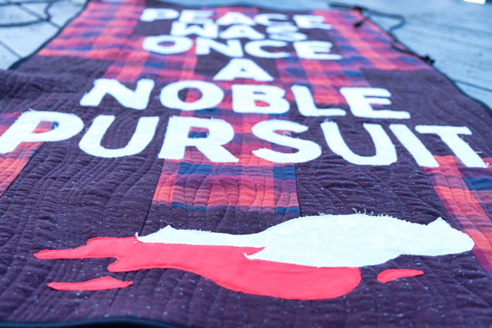 Close-up detail shot of the quilted textile banner showing the white text 'NOBLE PURSUIT' on the red and black plaid background, with visible quilting stitches and part of the white dove design at the bottom.