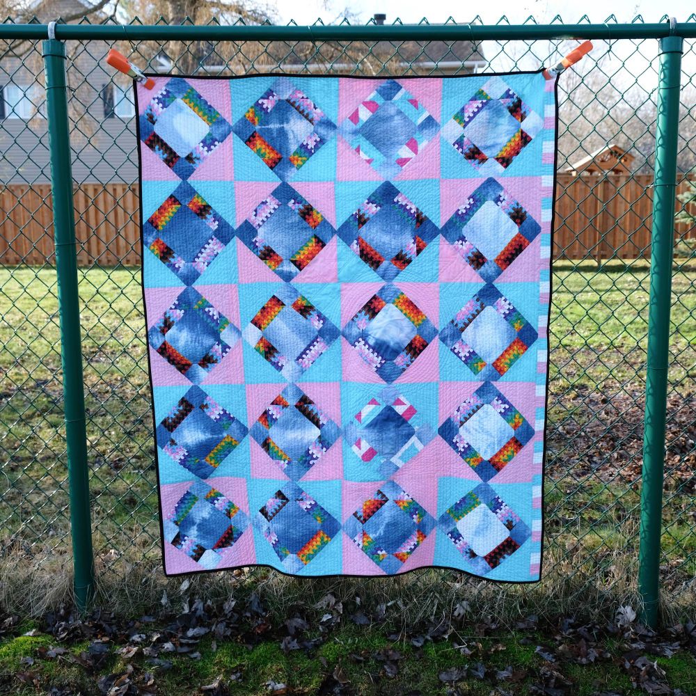 A detailed geometric quilt pattern featuring diamond shapes arranged in a 5x4 grid. The diamonds contain indigo-dyed fabric with rainbow accent pieces, set against alternating light blue and pink squares. The overall design creates a cohesive pattern that incorporates both traditional quilting techniques and pride flag colors. The quilt is displayed on the same chain-link fence with a black binding and orange clips holding it up.
