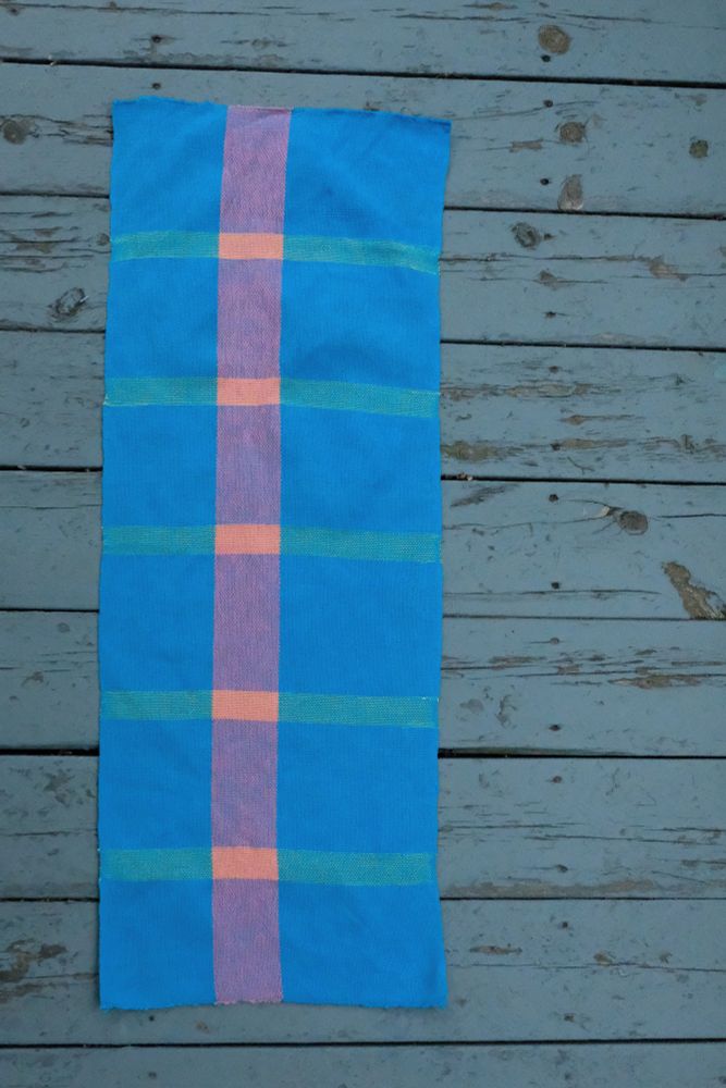 Bright blue rectangular handwoven textile with a central vertical pink stripe and horizontal teal lines forming a simple grid pattern, displayed flat on weathered blue wooden planks.