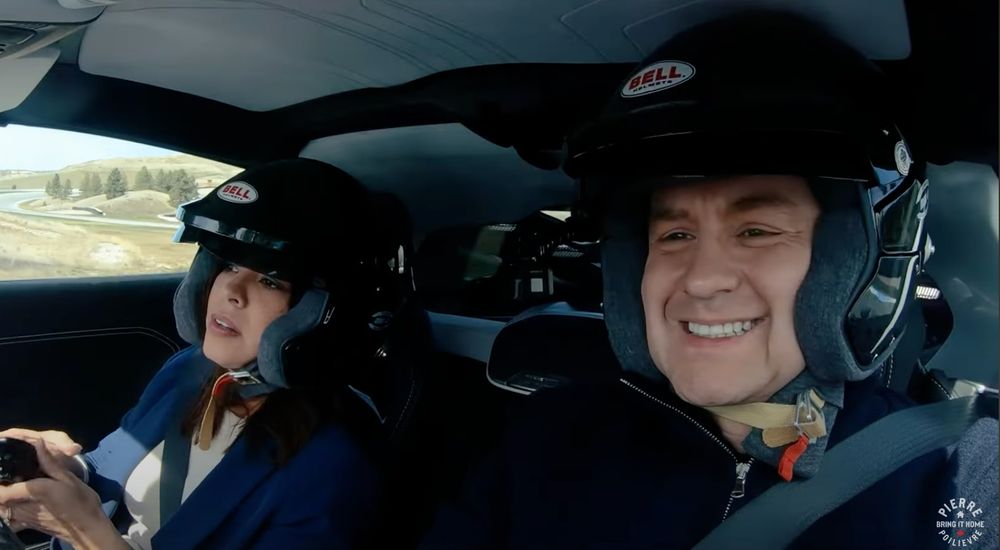The image shows two people in a car wearing Bell racing helmets - with a distinctly dorky vibe. The person on the right is smiling widely while the person on the left appears to be concentrating or reacting to the drive. They're inside what seems to be a performance or sports car, and the landscape visible through the window suggests they're on a racetrack or driving course. The image appears to be from a car entertainment or review show, with a "Pierre Poilievre" watermark visible in the corner. The staged nature of the photo op gives it an awkward, try-hard quality.

