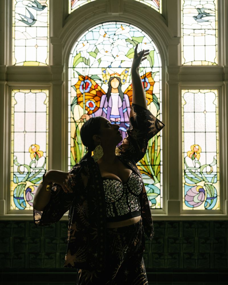 Bollyvvitch, a South Asian performer, stands in a dancing pose in front of an old stained glass window. 