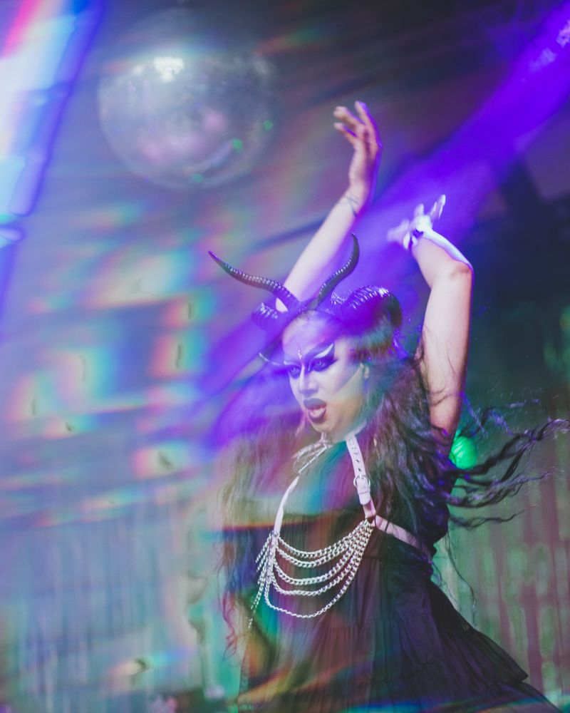 Bollyvvitch performs on stage, a disco ball above her and lens flares cascading across the photograph. She wears a black dress and horns, and pink body belt. Her hair is in pigtails, flowing wildly. Her arms are raised above her head, mid performance.