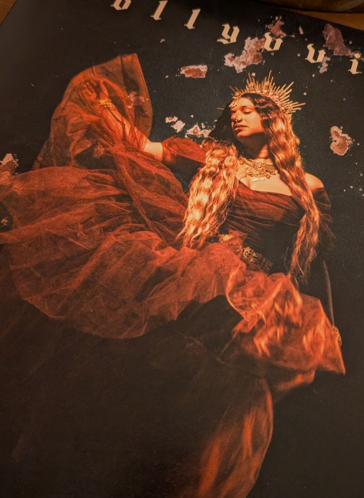 Album cover for As Above, So Below by Bollyvvitch. She stands in front of a dark background, dressed in a brown tulle gown and long dirty blonde hair. Her face is turned to the side, her eyes closed, wearing Indian gold jewellery and crown. Her arm is raised, lifting the dress up to look like she's floating.