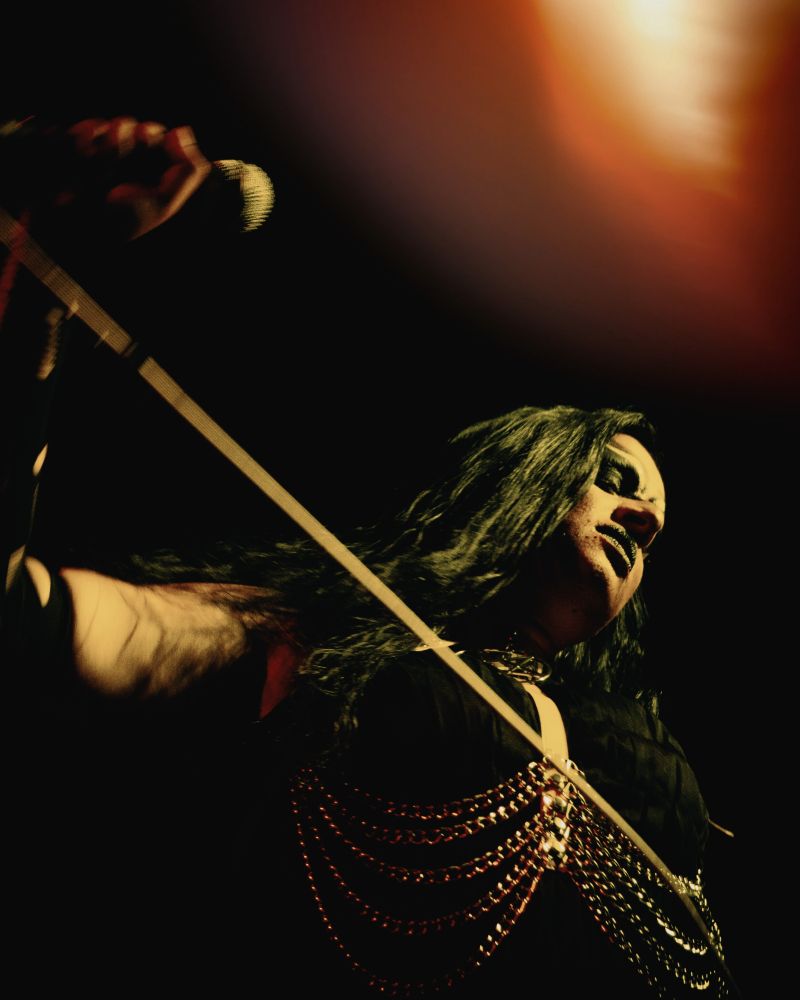 Bollyvvitch stands on stage wearing a black dress with a pink harness and chains. She holds the microphone and is looking down. A light leak in the corner illuminates her