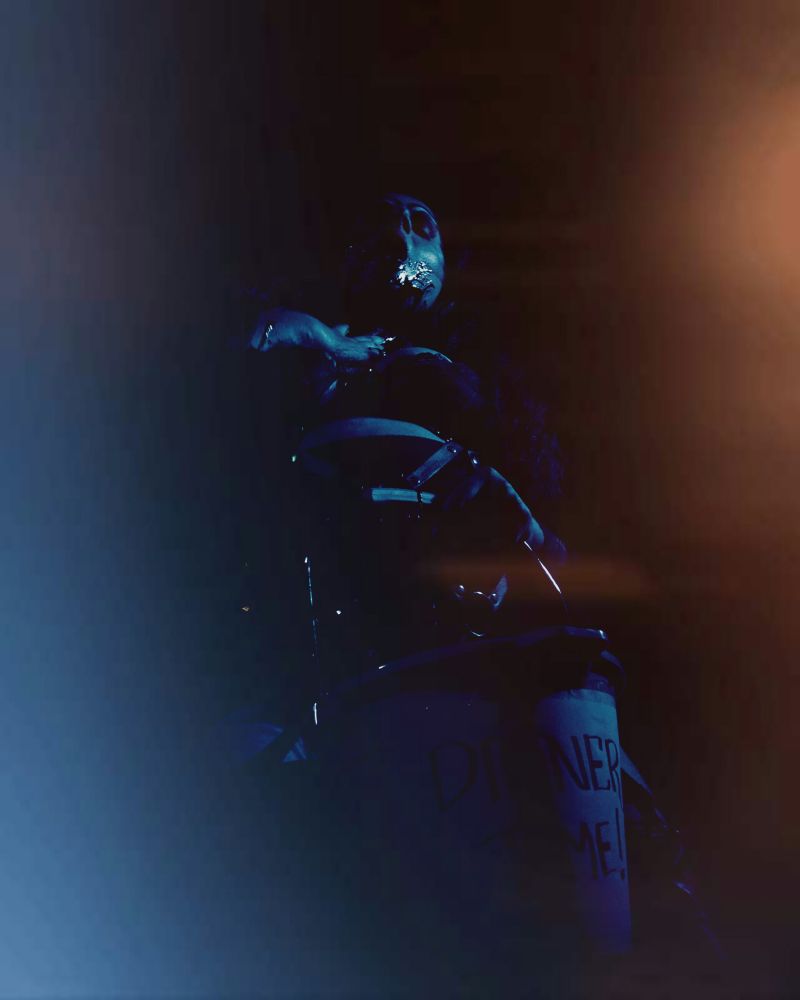 Bollyvvitch stands on stage wearing a gothic bolero and bra, with a black corset shaper and belt. The camera is at a low angle as she is smearing blood on herself, as she holds a bucket that says 'DINNER TIME' on the front. The colour tones are very dark, with some light leaks at the side. 