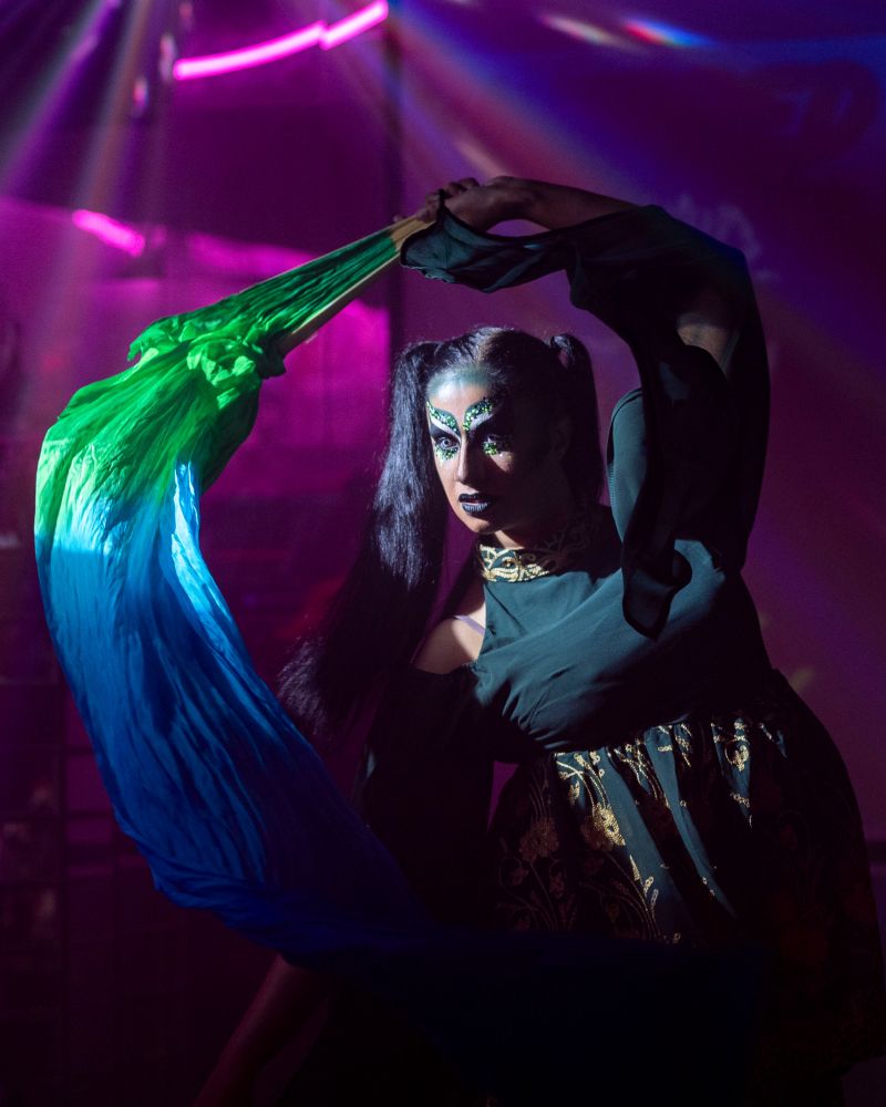 Bollyvvitch is mid-performance in a dimly lit room of purple lights. A white spotlight is on her, showing her green snake-like make up and green dress. She holds a green and blue veiled fan over her head.