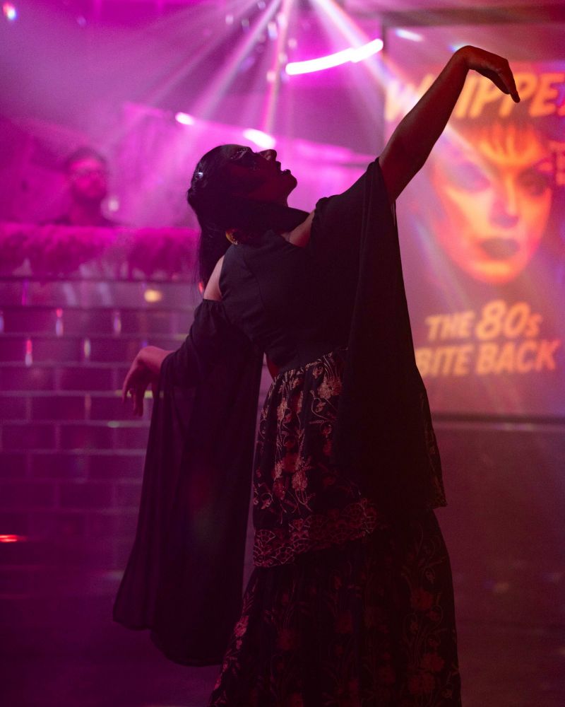 Bollyvvitch stands mid performance, with glowing lights behind her. Her silhouette is of her standing, her head tilted up with one hand raised like a snake head. 