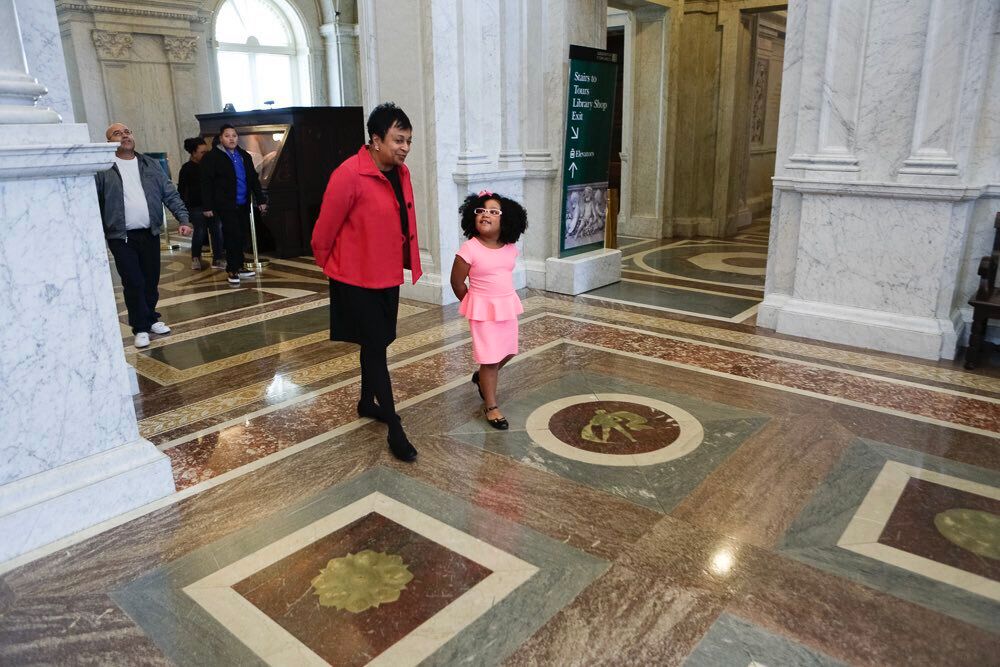 Four-year-old child Daliyah Maria Arana in a pink dress with matching pink hair bow walking with Librarian of Congress Carla Hayden inside the Library of Congress building. Three members of the child’s family are in the background, looking up at the building interior. Photo from Librarian of Congress Twitter account.