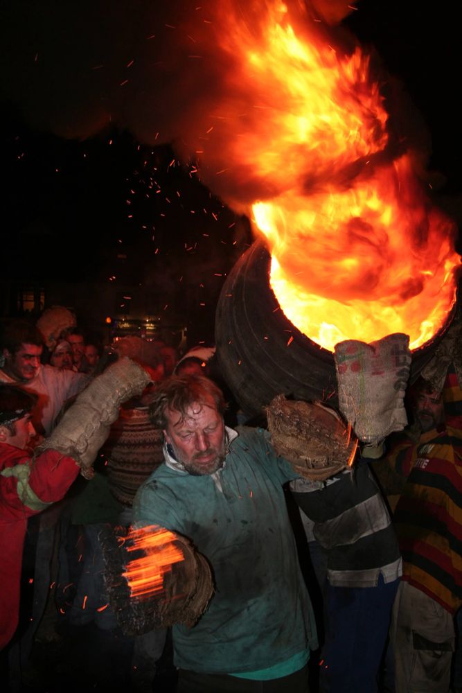 A man runs through the streets of Ottery St. Mary with a flaming tar barrel on his back. Another man, with soot on his face, waves his burning sack-cloth glove to try an extinguish the flames. Sparks fly into the night sky.