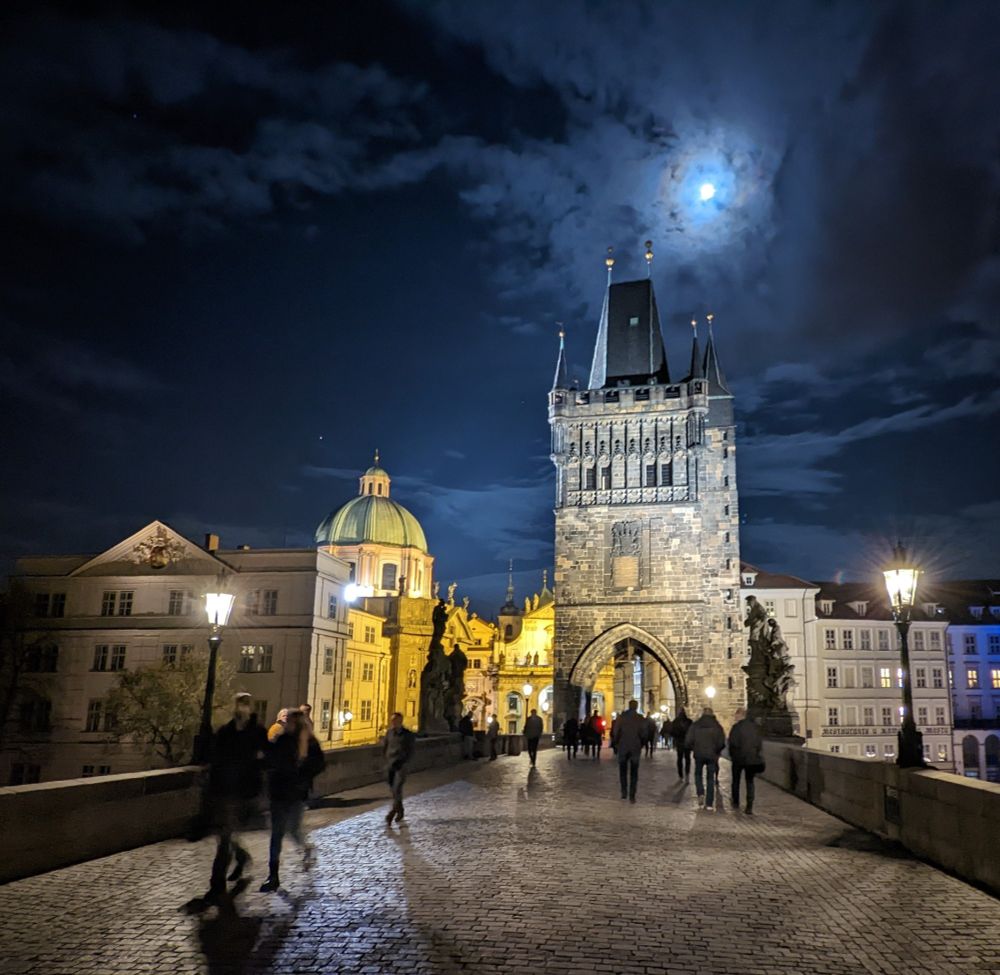 Charles Bridge at night. The moon is high in the sky filtered by thin clouds. The buildings are illuminated by warm, yellow light, contrasting with the deep blue of the night sky.