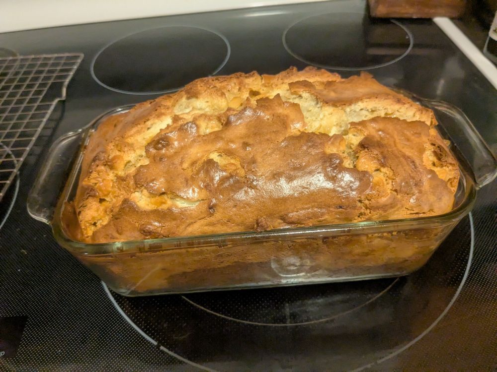 A fresh loaf of banana bread. The bread is still in a glass loaf pan on top of a stove top. The crust is a dark brown with some lighter colors showing through the cracked top of the bread.