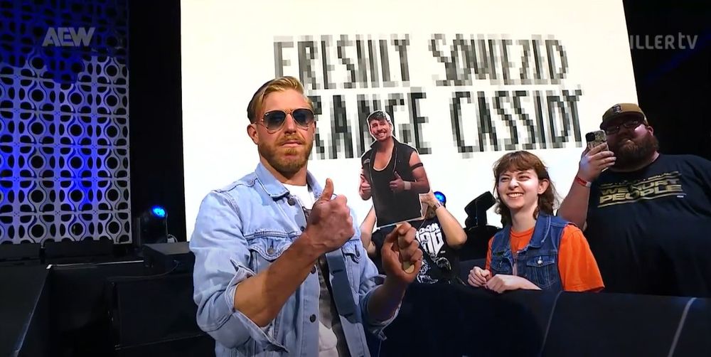 orange cassidy posing with a cut out of chuck taylor on a stick while the individual who brought it is smiling and thoroughly enjoying the moment. 
