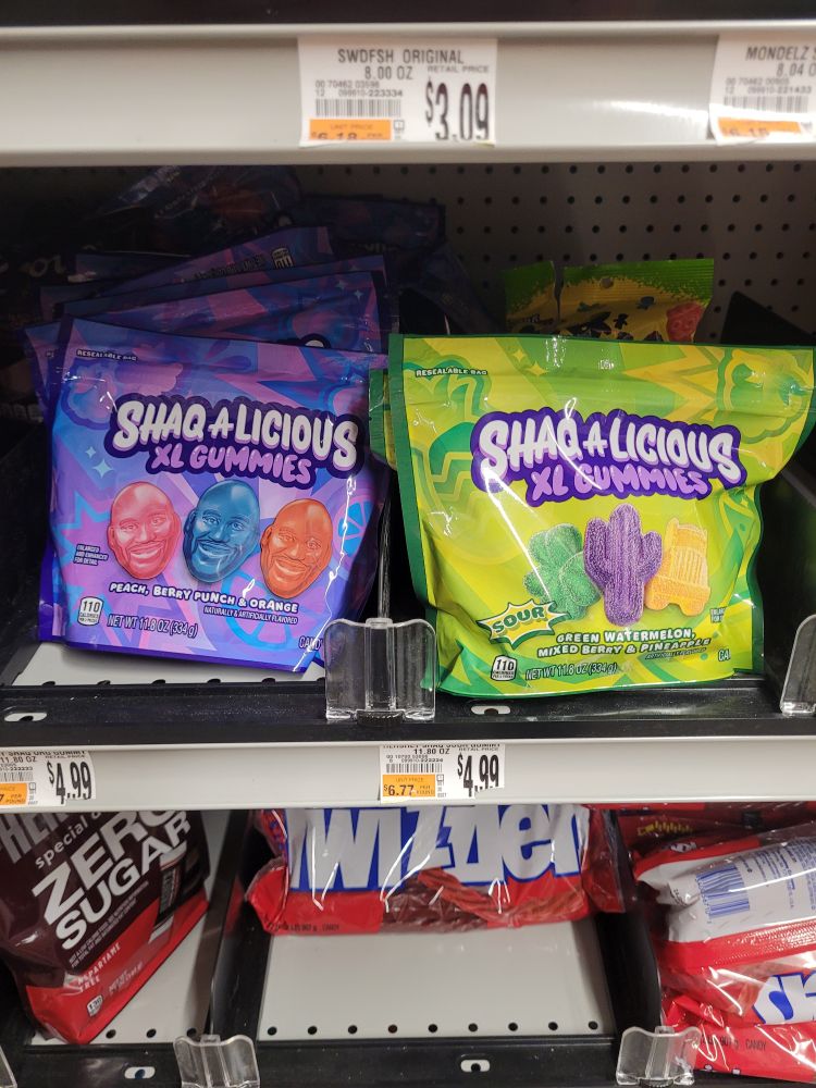 2 packages of shaq-a-licious xl gummies. the left bag is purple and features pink, blue, and orange gummie shaq faces. the right bag is green and the gummies are sour and shaped like a shamrock, a cactus, and a big rig