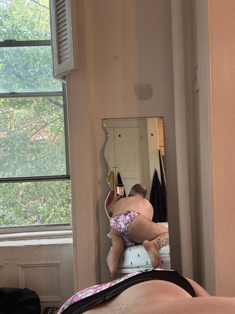 A man taking a selfie in a mirror bent over his bed in a pink and black speedo printed with Tom of Finland cartoons