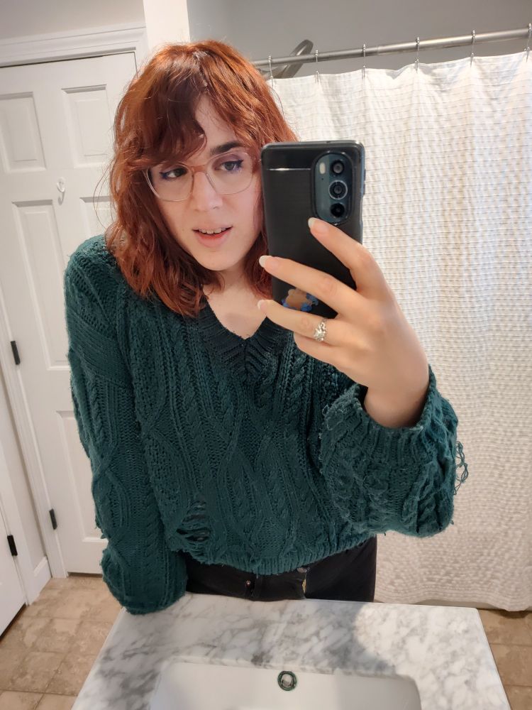 mirror selfie of me in a bathroom. my mouth is partially open, smirking. I'm leaning forward slightly on the sink. I'm wearing a dark green knit sweater, black pants, and transparent pink glasses. I have a silver ring on one finger 