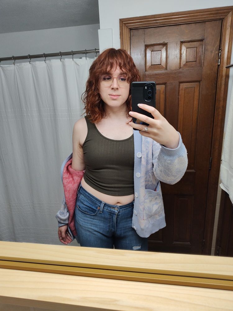 mirror selfie of me in a bathroom. I'm wearing jeans, a green crop tank top, and a white and blue cardigan. my hair is copper red and im smirking a bit. 