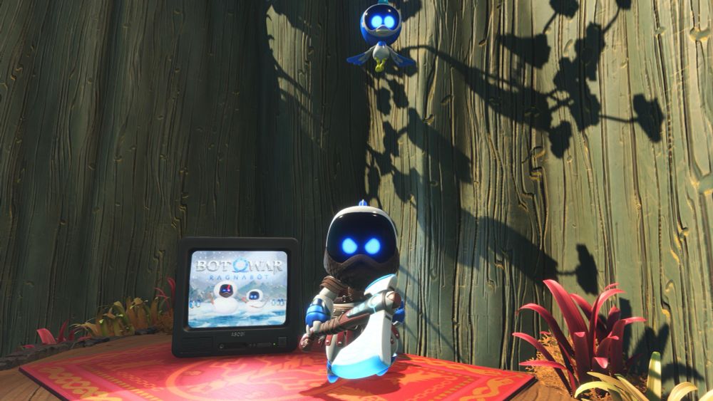 Astro bot posing in front of a tv with a fictional “Bot of War” game on the screen. Astro bot is also wearing an outfit similar to the one Kratos wore in the God of War Ragnarok video game. 