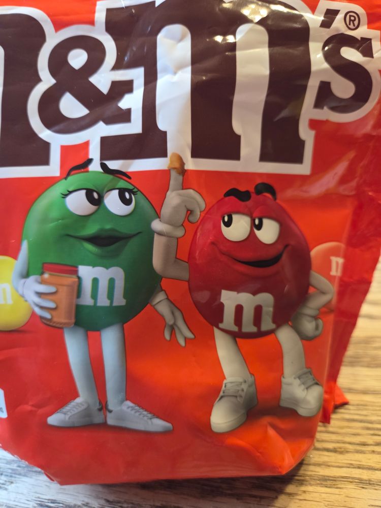 Package of peanut butter M&Ms with two cartoon M&Ms with one holding up a finger with peanut butter on it