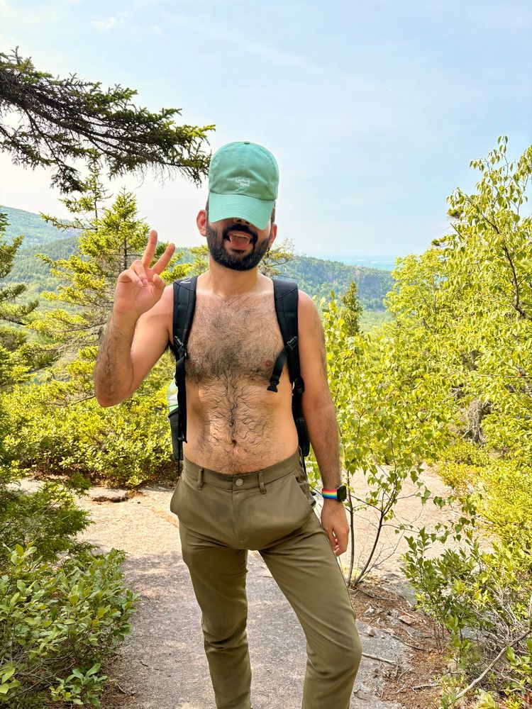 A person with a beard stands in a green, outdoor setting, wearing a light green cap and dark pants. They are shirtless, giving a peace sign while sticking out their tongue, with trees and a distant landscape visible behind them.