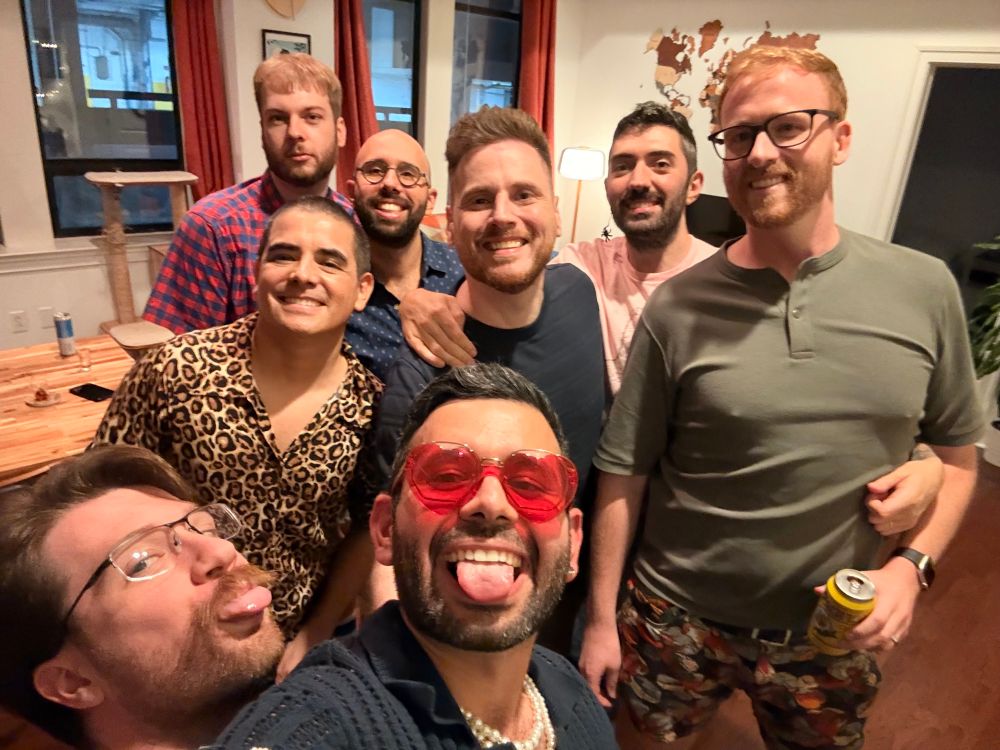 A group of eight friends posing together in a casual indoor setting, smiling and making playful faces. The atmosphere appears joyful, with various clothing styles, including a leopard print shirt and colorful shorts.