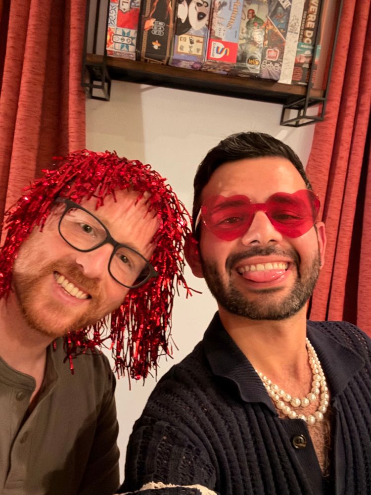 Two men are posing for a selfie. One wears a bright red, shiny wig and glasses, while the other sports heart-shaped pink sunglasses and multiple pearl necklaces. The background features a shelf with board games and a curtain. Both are smiling and displaying playful