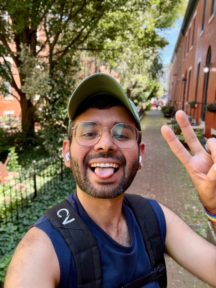 A person is taking a selfie outdoors, smiling and sticking out their tongue. They are wearing a green cap and glasses, with wireless earbuds in their ears. The background features a lush, leafy environment with red brick buildings and a pathway lined with greenery.