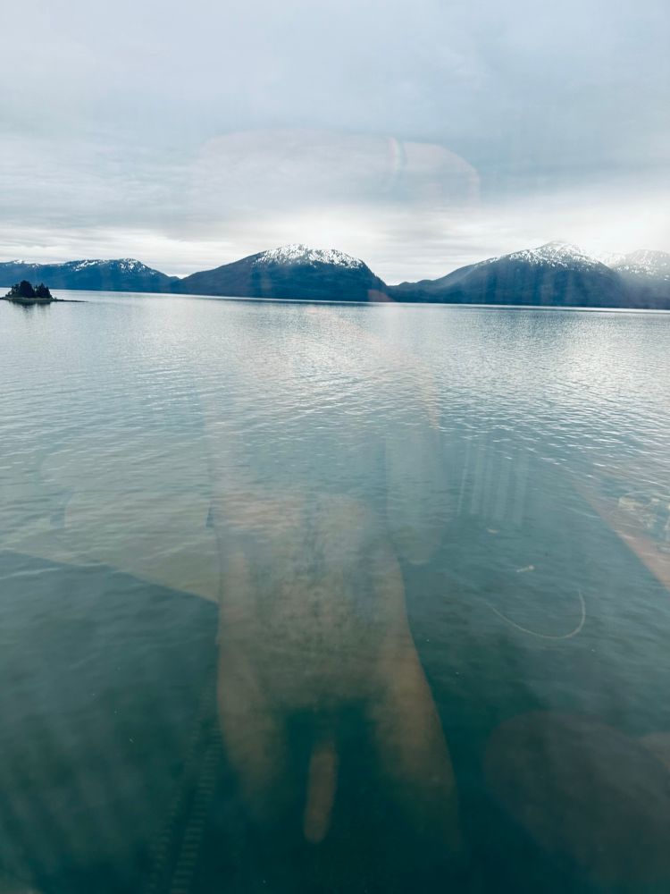 A tranquil body of water reflects snowy mountains under a cloudy sky, with an island visible in the distance. There appears to be a reflection in the water that adds a layer of complexity to the scene.