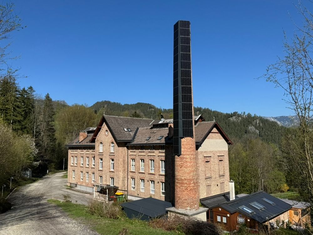An old brick building with a smokestack. The smokestack is now covered with PV panels. 